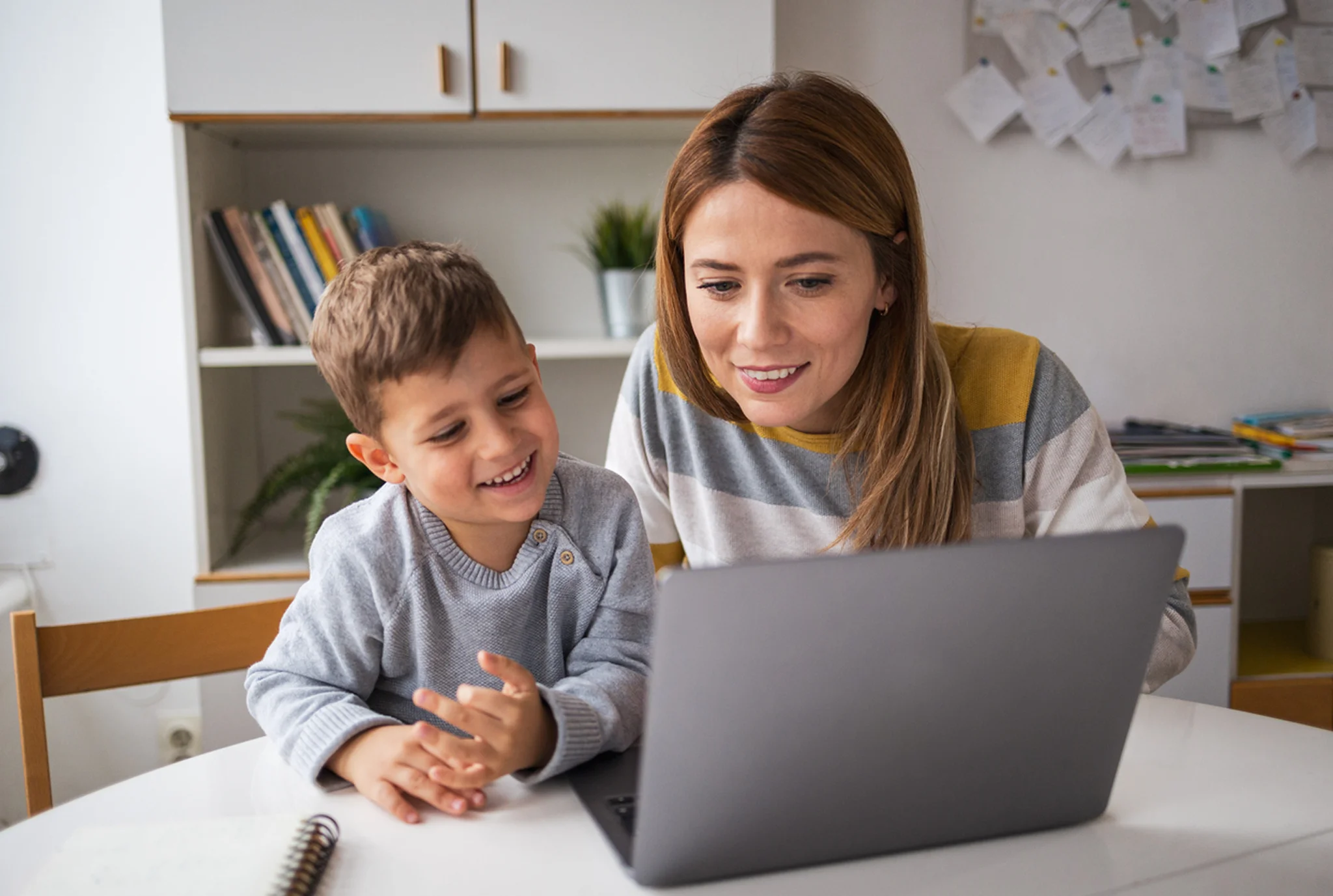 A woman and a young boy smile while looking at a laptop screen in a cozy room. Books and papers are on the shelves and walls, creating a warm, learning environment.