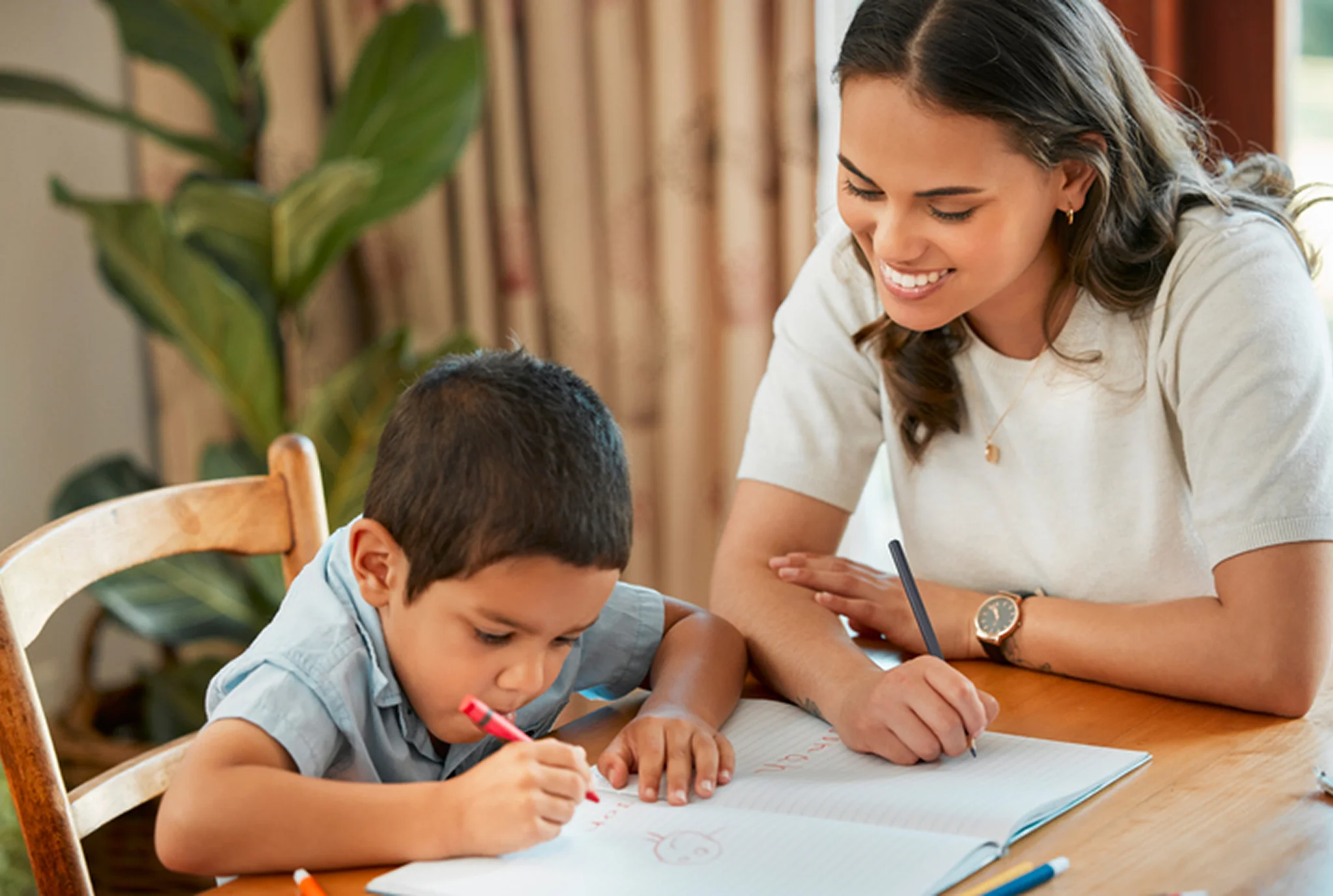 A smiling woman supports a young boy drawing with a red pencil at a wooden table, conveying a warm, educational, and nurturing atmosphere.