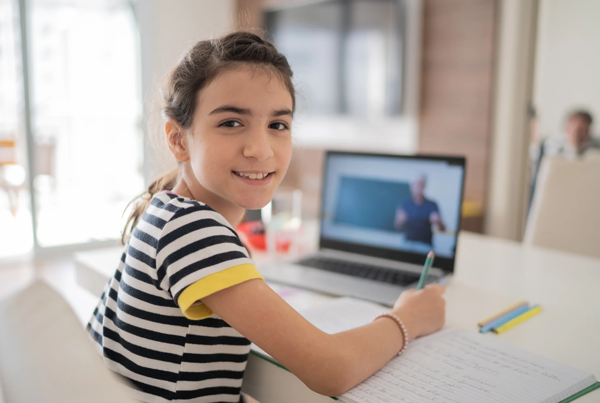 A young girl in a striped shirt smiles while studying at a white table. She's writing in a notebook, with a laptop screen showing an online lesson.
