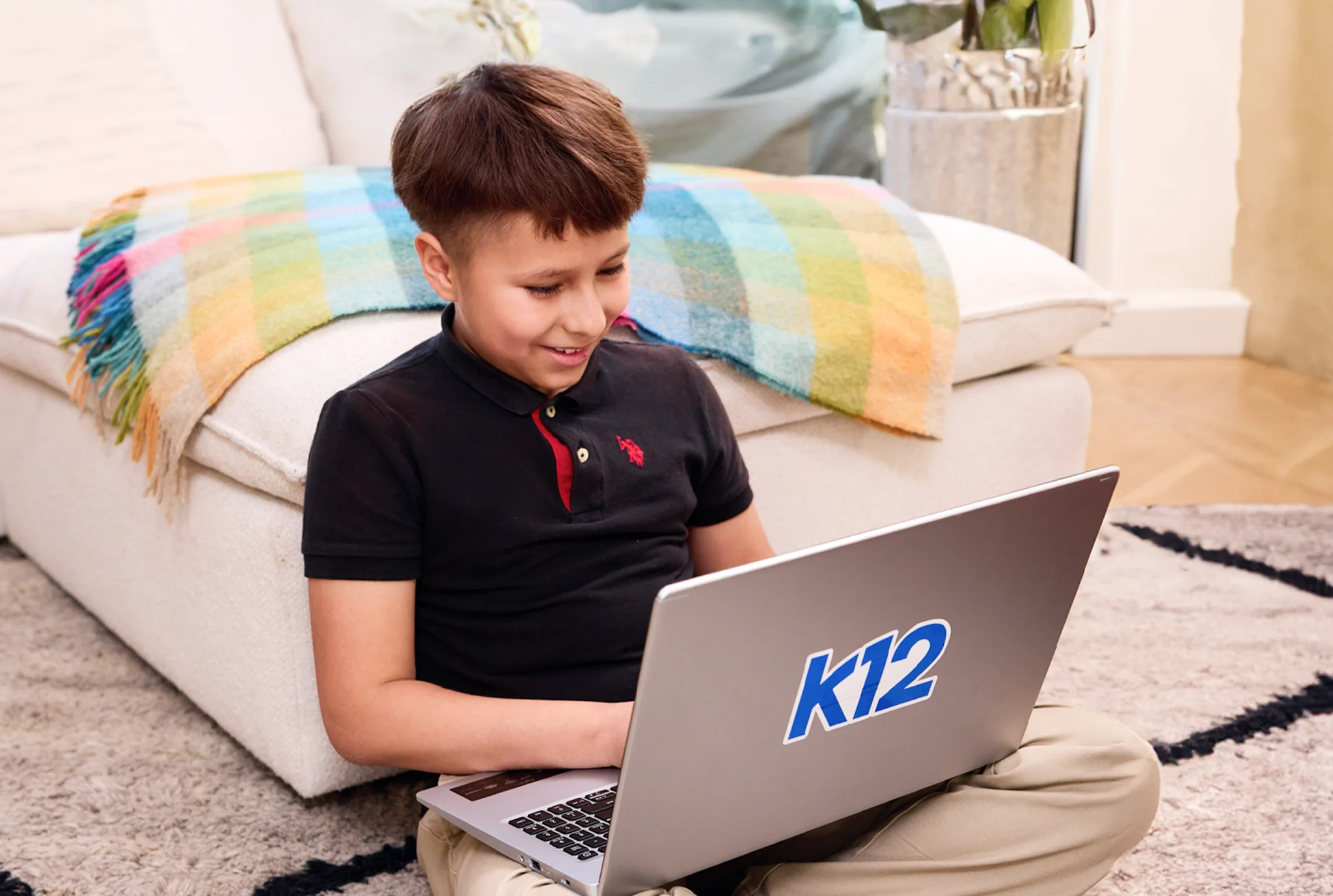 Young boy sitting cross-legged on a carpet, smiling at a laptop with a "k12" sticker. A colorful blanket is draped over a sofa behind him.