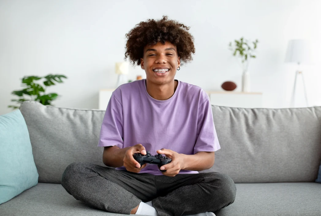 Young person with curly hair and a purple shirt, smiling while holding a game controller. They sit cross-legged on a sofa in a bright, cozy room.