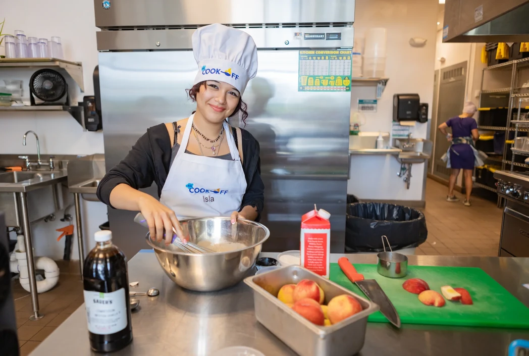 Young person smiling, wearing a chef's hat and apron, mixes ingredients in a bowl in a commercial kitchen. Apples and kitchen utensils are on the counter, conveying a cheerful culinary atmosphere.