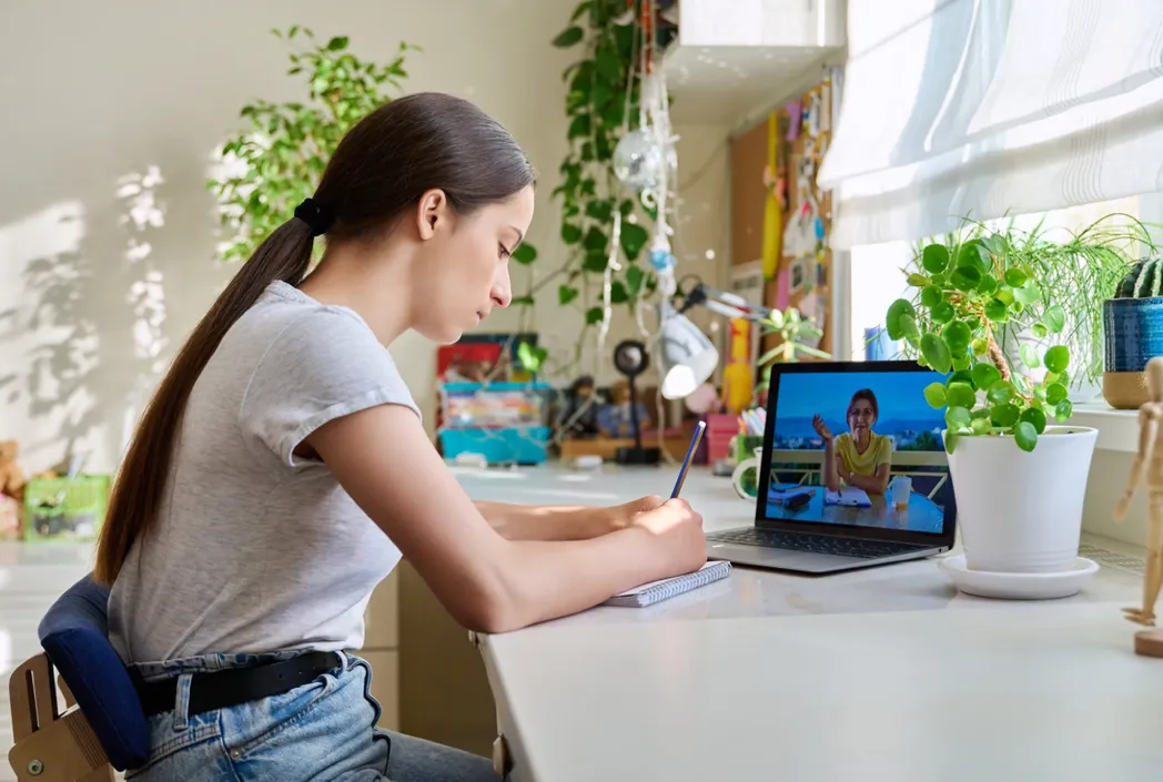 A young woman sits at a desk, writing in a notebook. A laptop with a video call is open beside her. The room has plants and a sunny ambiance.