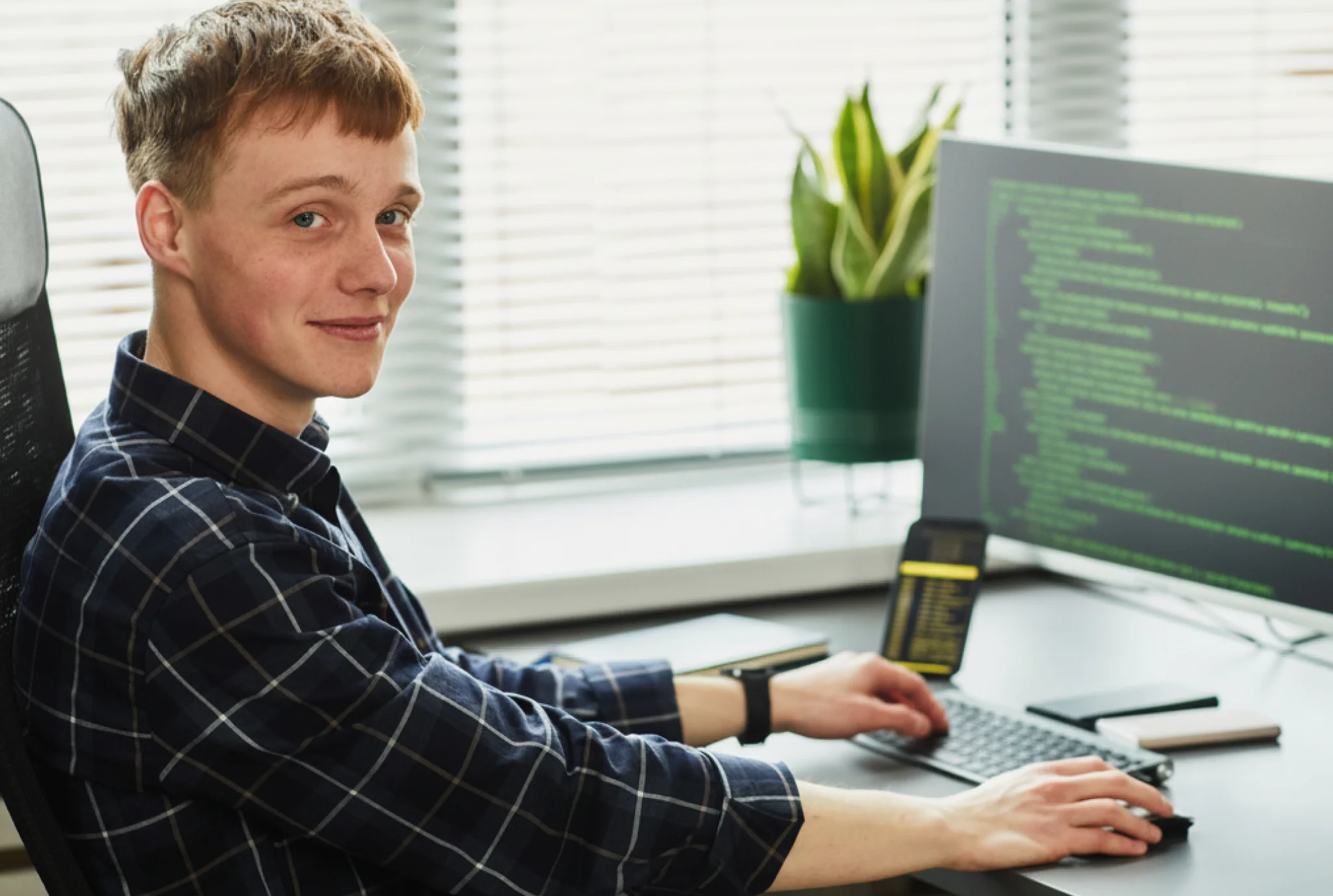Young man in a plaid shirt smiles while typing at a desk with a monitor displaying code. A potted plant and window blinds are in the background.