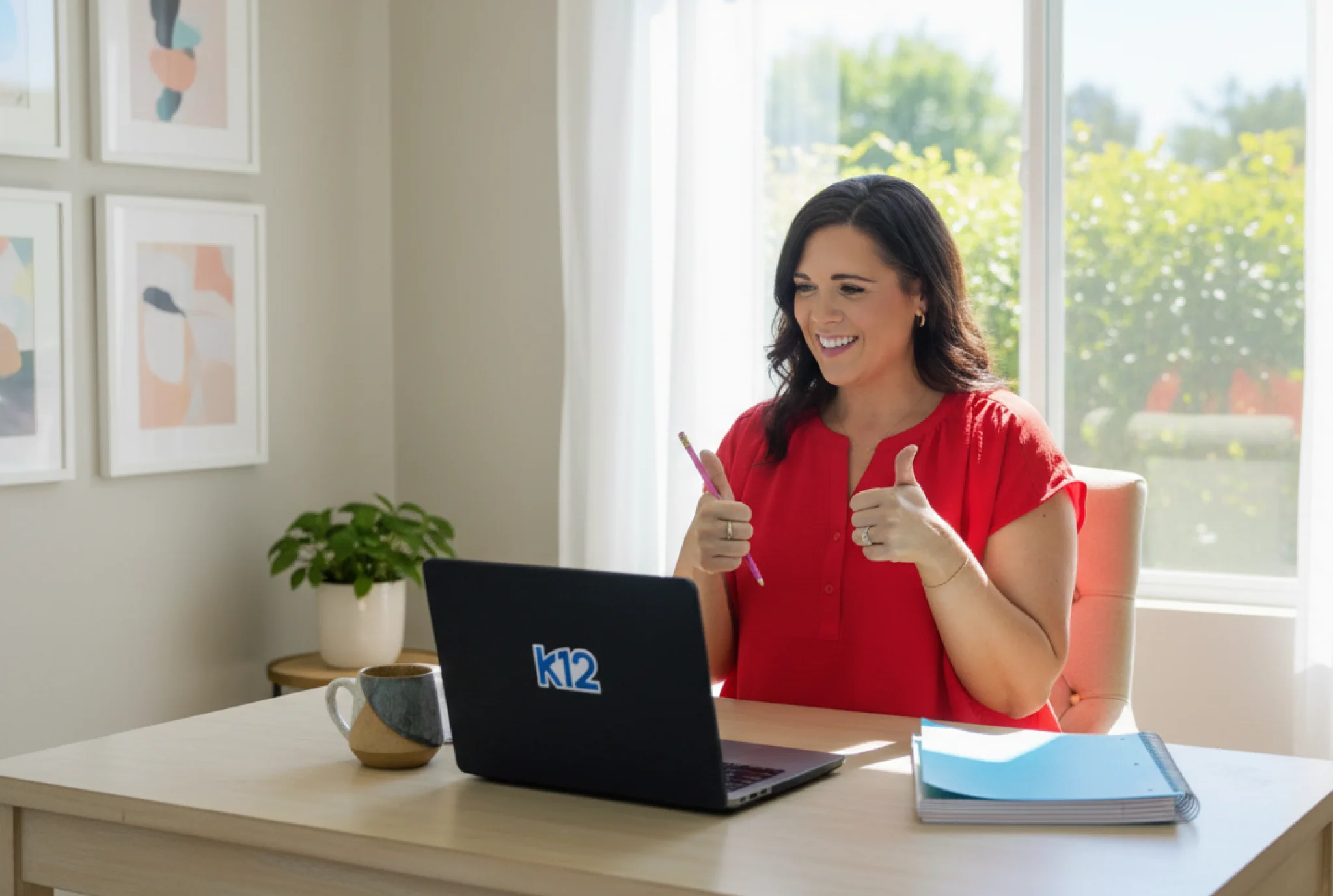 Smiling woman in a red shirt giving thumbs up during a video call on a k12 laptop. She's seated at a desk with a mug and notebooks, in a bright room.