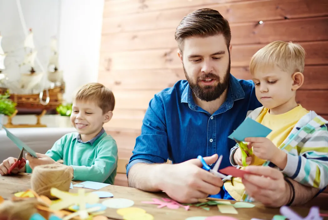 A father and two young boys are engaged in a paper craft activity at a table, surrounded by colorful cutouts. Everyone appears focused and joyful.