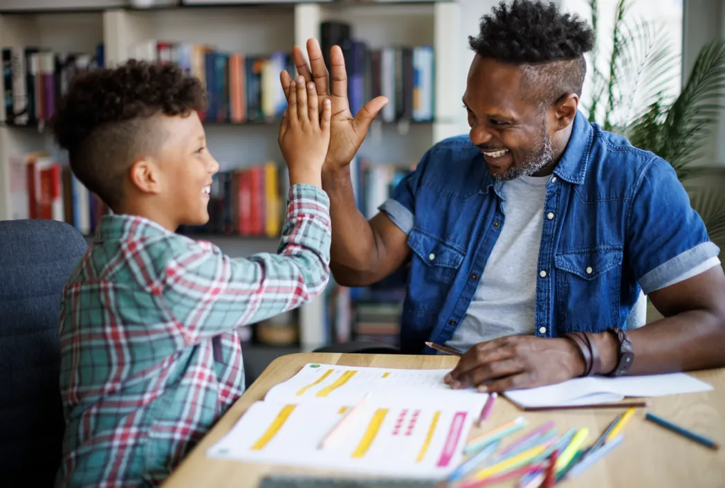 Father and child smiling and high-fiving at a desk with colorful papers and pencils, conveying a joyful and supportive learning environment. Bookshelves in background.