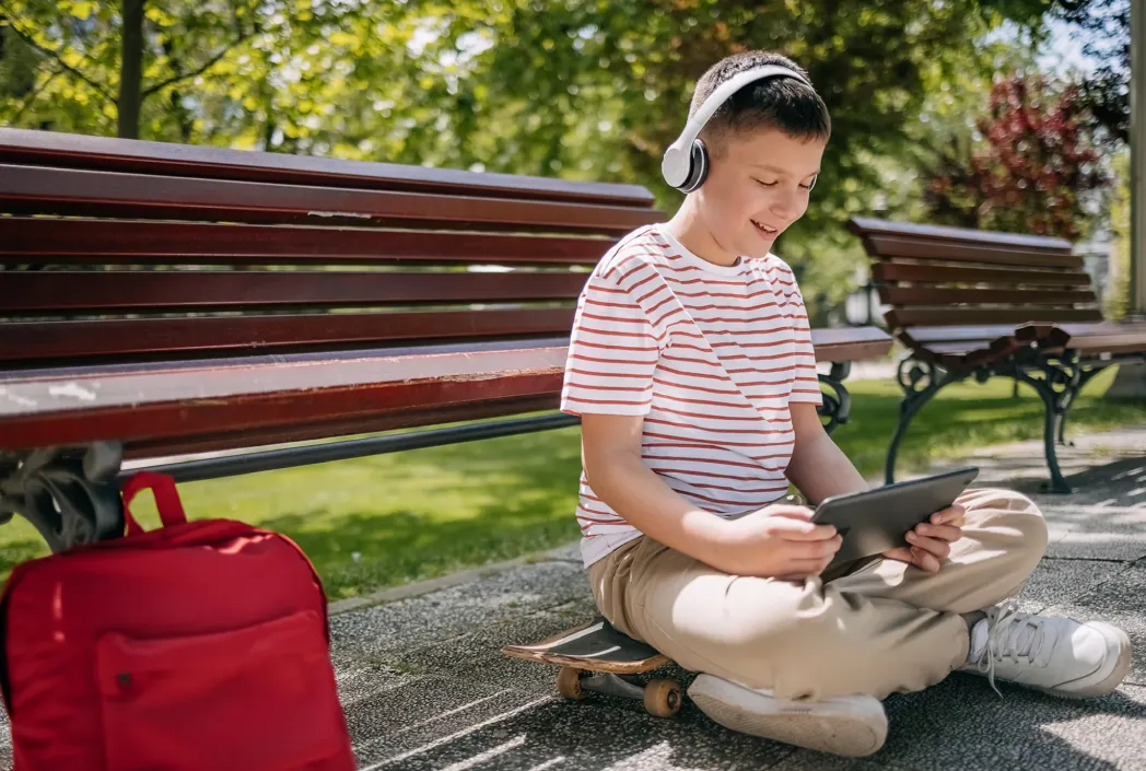 A boy wearing headphones and a striped shirt sits cross-legged on a skateboard, using a tablet. He's in a park next to a red backpack. Relaxed, sunny setting.