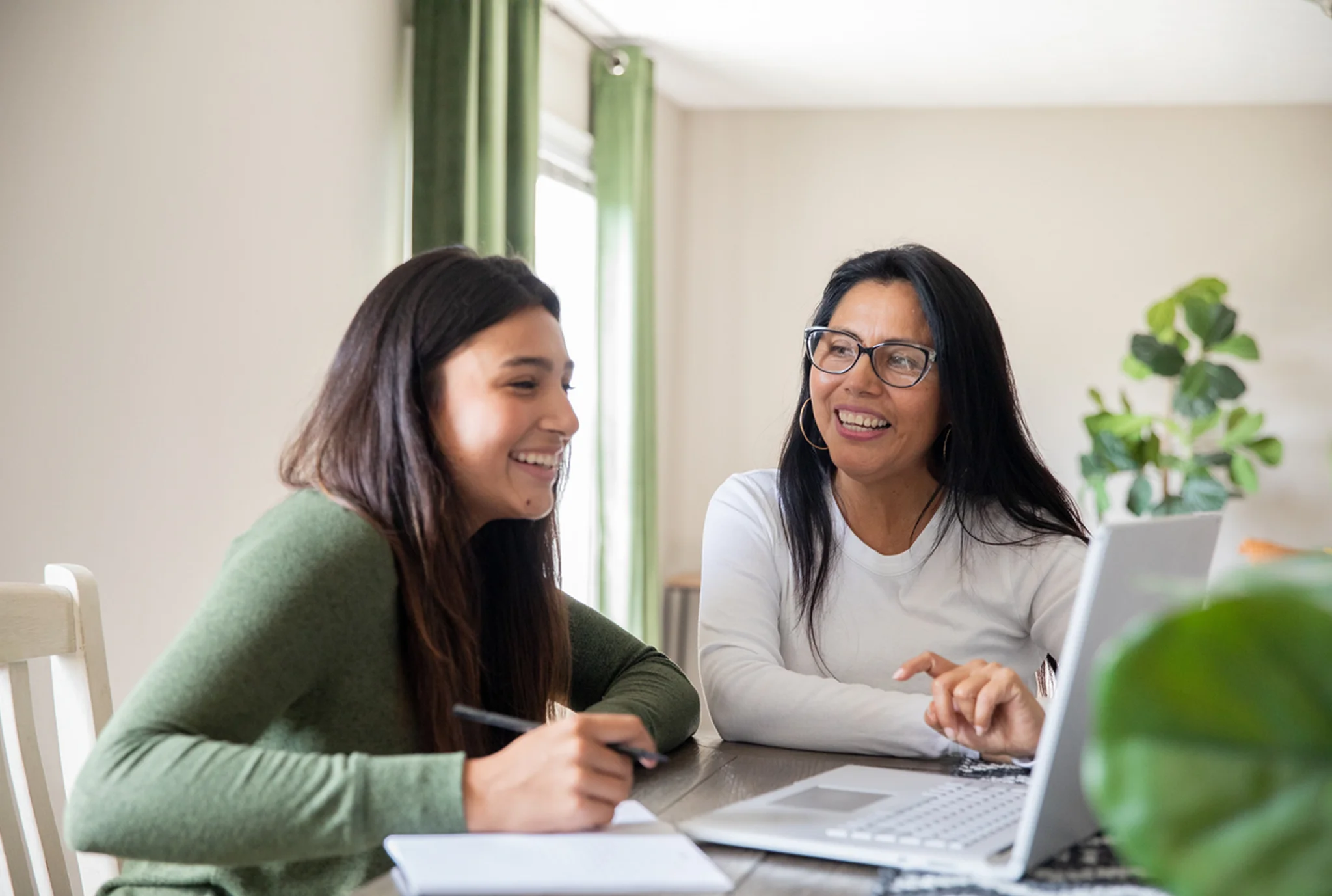A young boy wearing headphones waves at his laptop during an online class. He's seated at a desk with colorful pencils, books, and a globe nearby, projecting a cheerful mood.