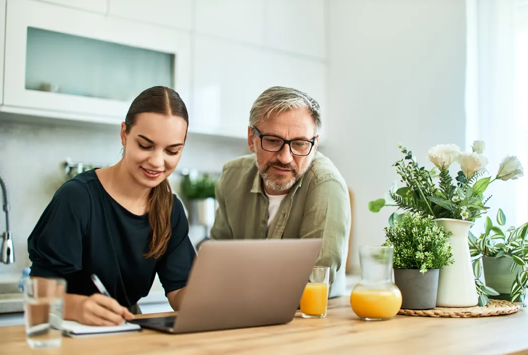 A woman and man are smiling at a laptop in a bright kitchen. The woman takes notes, while orange juice and potted plants sit on the wooden table.