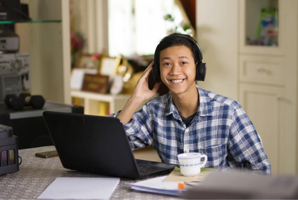 A smiling person in headphones sits at a table using a laptop. They wear a checkered shirt, and a cup and papers are on the table in a cozy room.