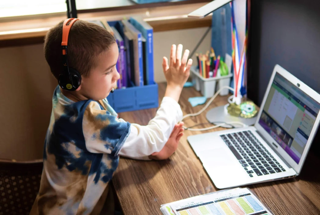 Young boy with headphones sits at a wooden desk, engaged with a laptop in a well-lit room. Books and stationery are neatly arranged nearby.