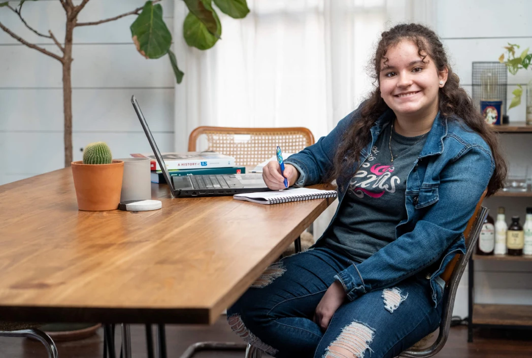 A young woman smiles while writing in a notebook at a wooden table with a laptop, books, and a cactus plant. The room is cozy and well-lit.