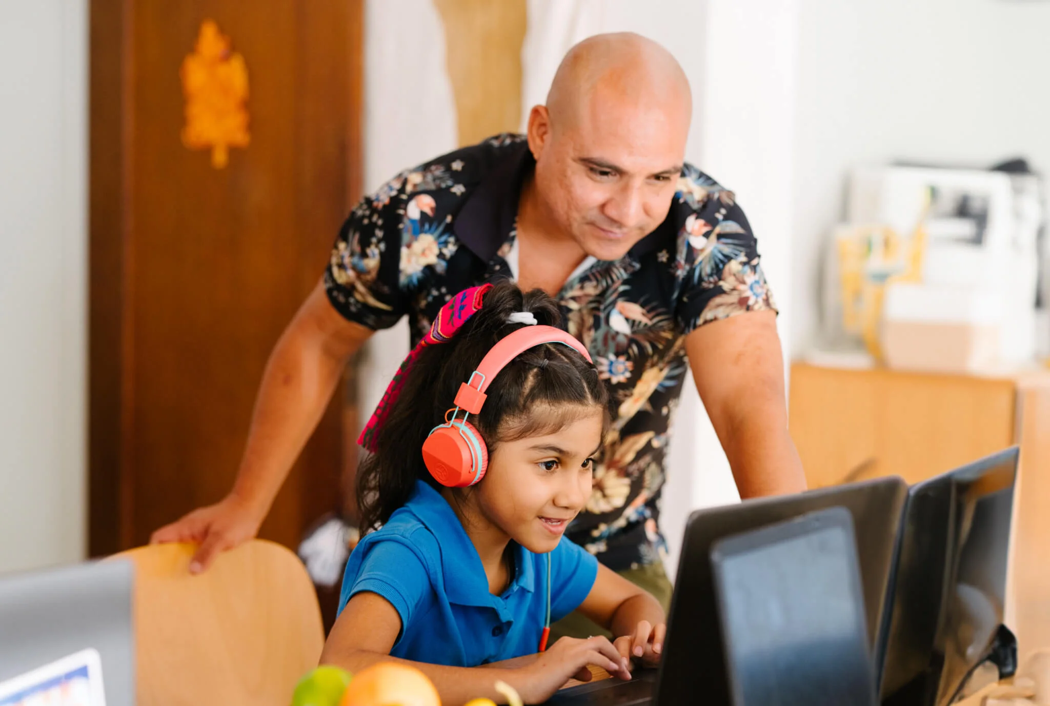A smiling girl wearing red headphones sits at a desk using a computer, while a man in a floral shirt leans over, watching supportively.