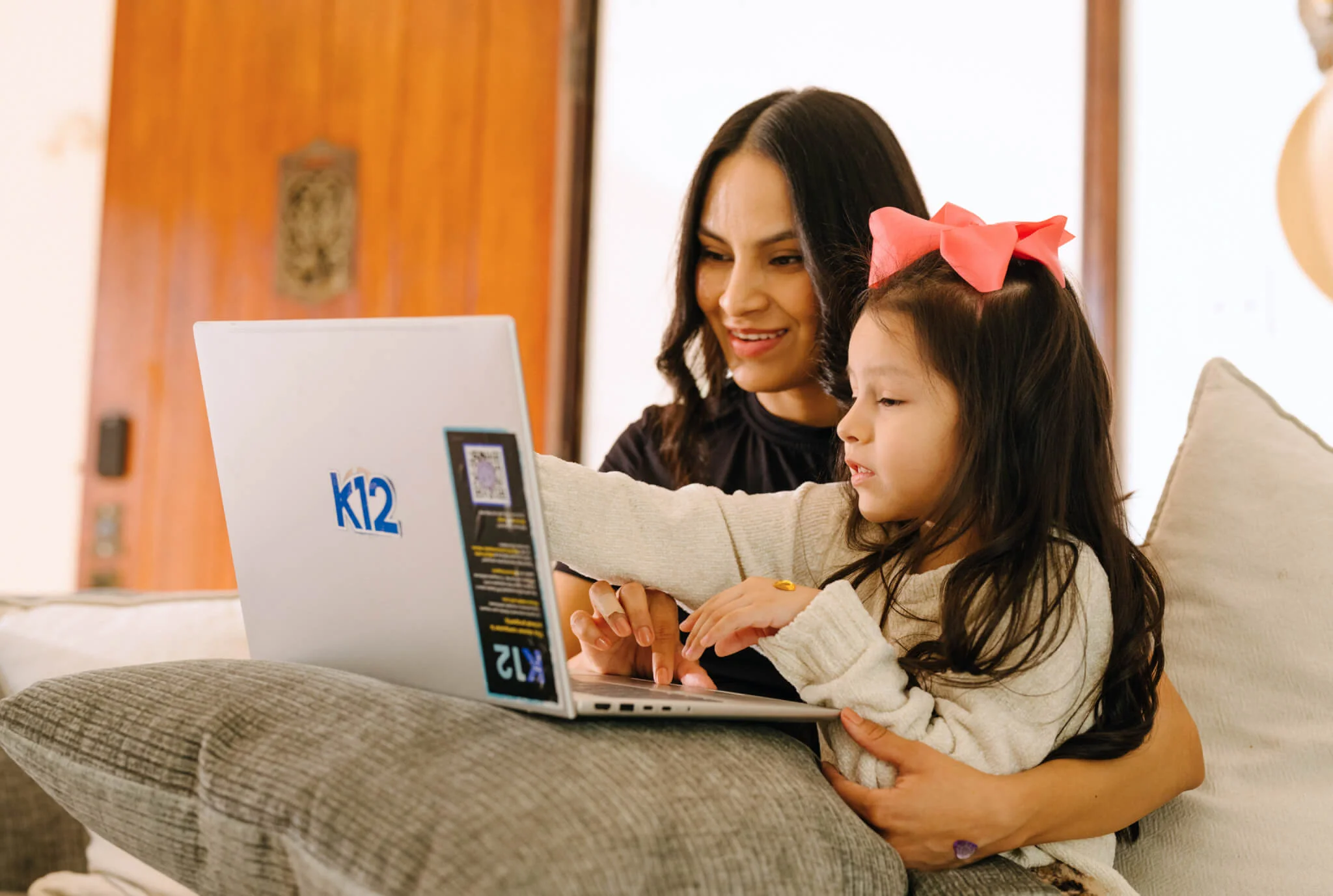 A woman and young girl with a pink bow sit on a couch using a laptop with "k12" on it. They look engaged and happy, sharing a learning moment.