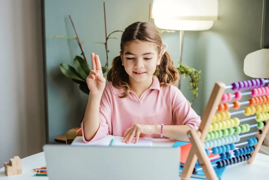 A young girl with brown hair in pigtails sits at a desk, smiling and raising two fingers. She uses a colorful abacus, surrounded by plants and soft lighting.