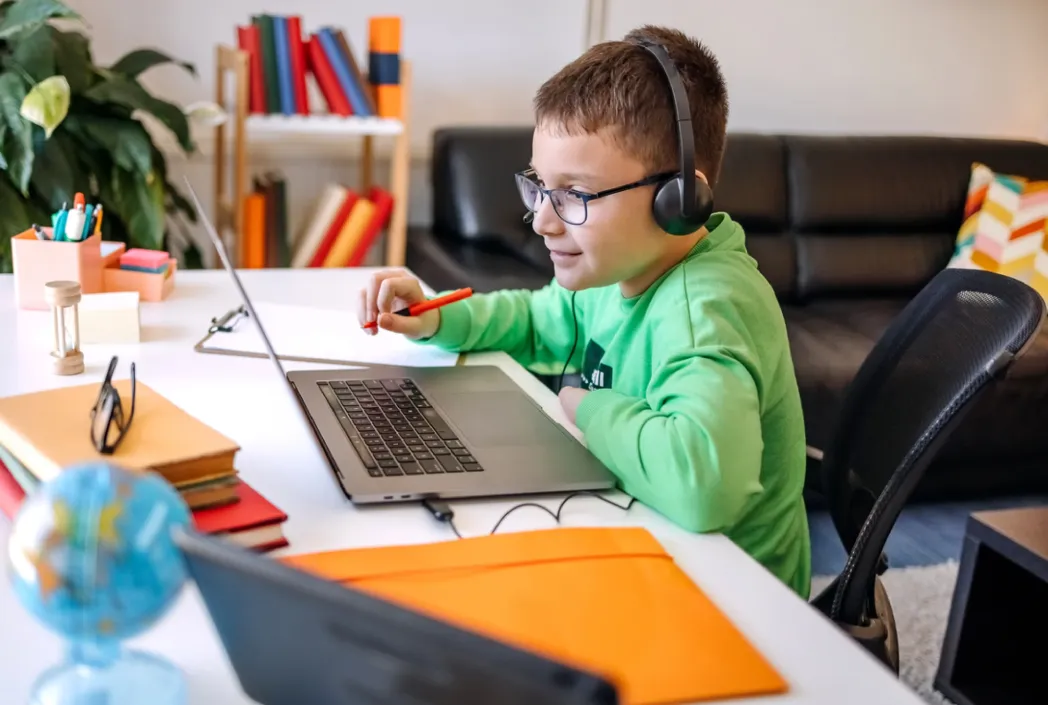 A boy in glasses and headphones, wearing a green shirt, studies intently at a laptop on a white desk. Books, a globe, and a sofa are in the background.