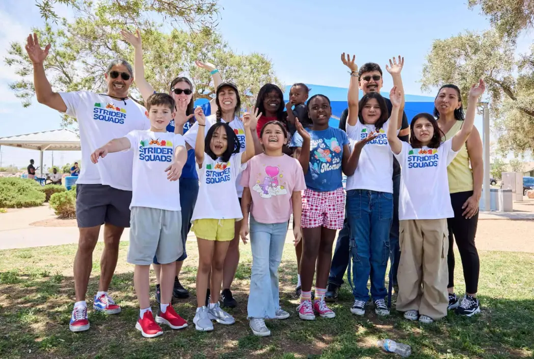 A diverse group of smiling people, adults and children, pose outdoors with arms raised under trees. They all wear "Strider Squads" shirts, exuding joy.