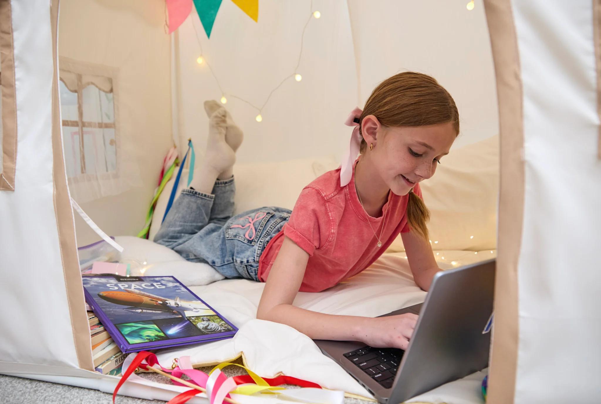 A young girl is lying on her stomach in a tent, smiling and typing on a laptop. She is surrounded by colorful books and festive bunting, creating a cozy atmosphere.