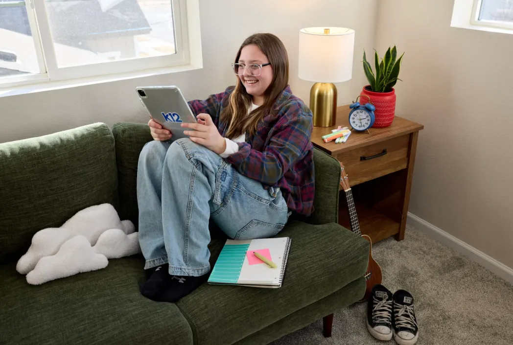 A smiling person with glasses sits on a green couch, holding a tablet labeled "K12." Nearby are a notebook, lamp, clock, and guitar, conveying a cozy, studious vibe.
