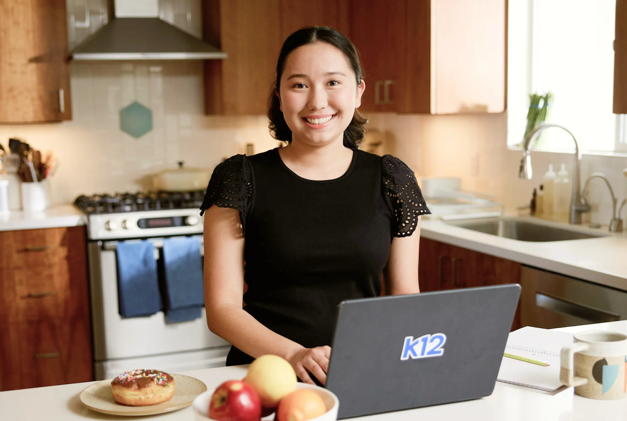 A woman stands smiling in a kitchen using a laptop with "K12" on it. A plate with a donut and apples is in the foreground, creating a warm, inviting scene.