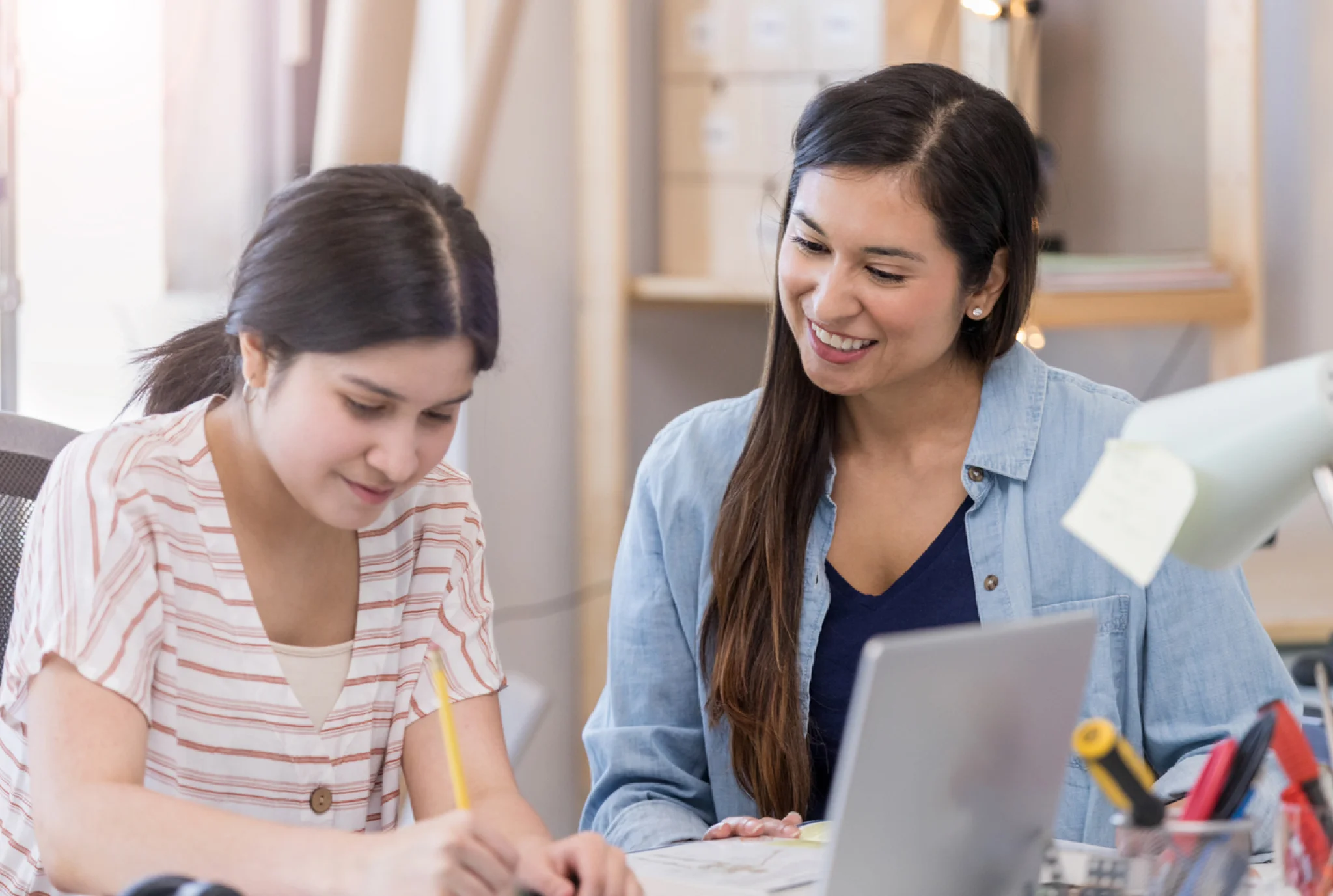 A woman smiles supportively as a younger girl writes at a desk, suggesting a tutoring or mentoring scene. A laptop and study materials are visible.