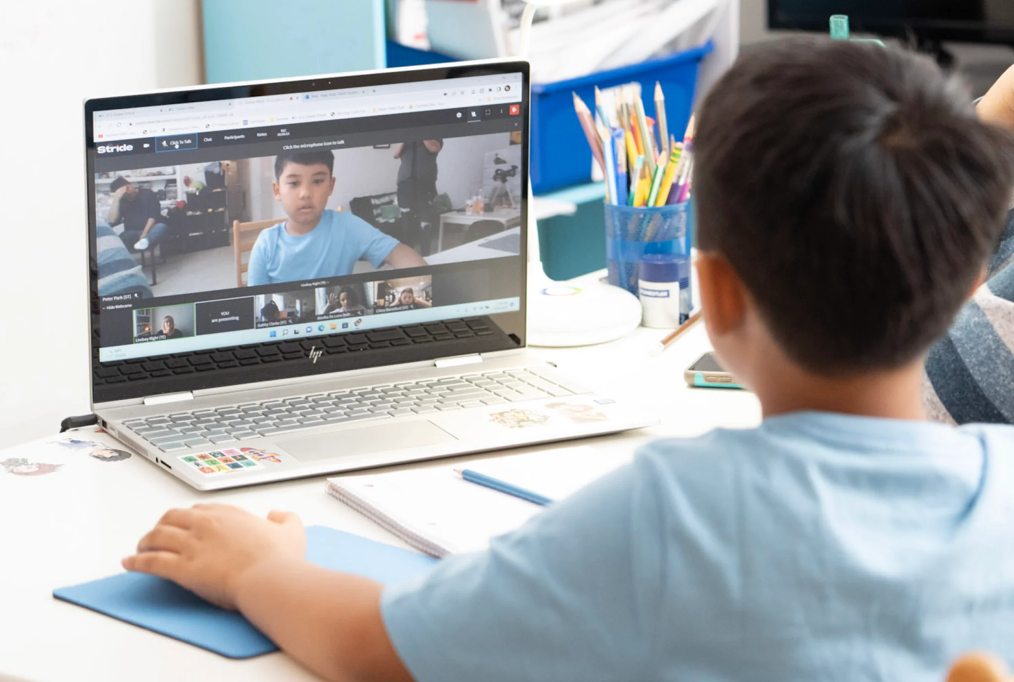 A young boy in a blue shirt sits at a desk, participating in an online class on a laptop. The screen shows other classmates on a video call.