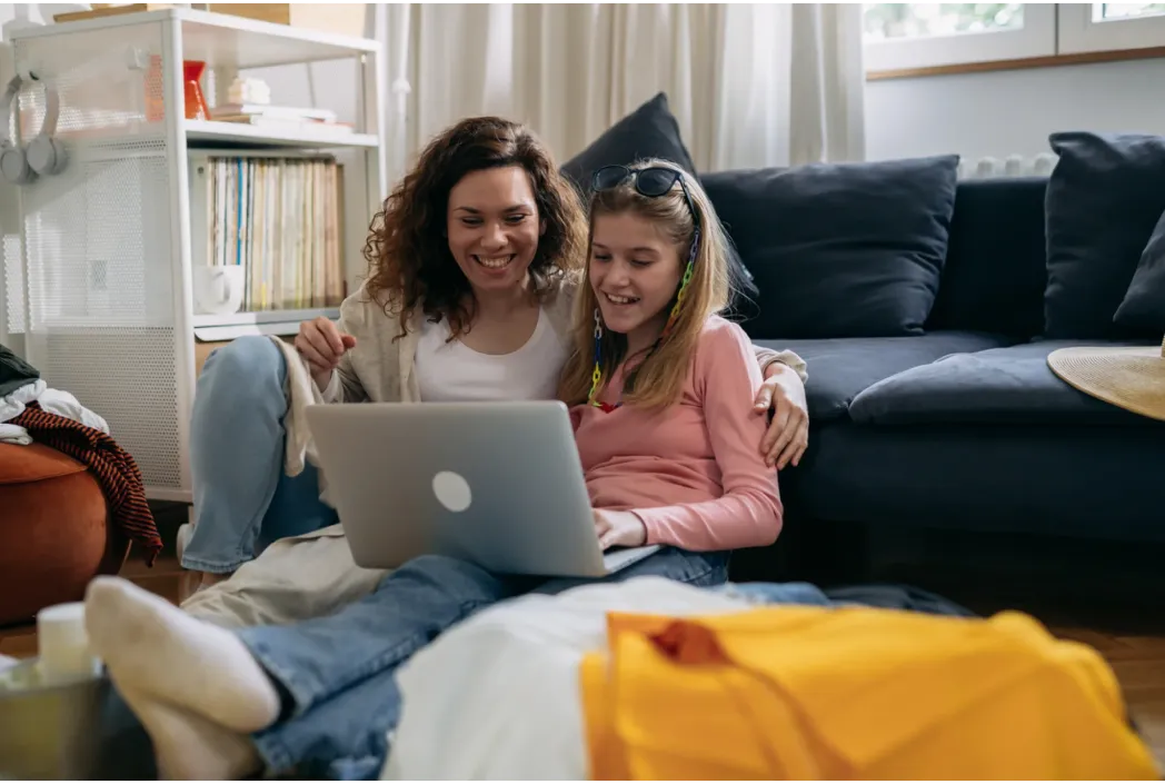 A woman and a young girl sit on the floor, smiling and looking at a laptop. They appear relaxed and happy, surrounded by homey, casual interior decor.