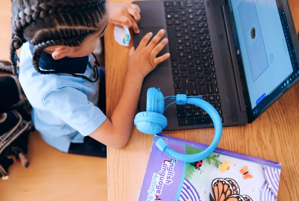 A young child with braided hair uses a laptop, focusing on the screen. Bright blue headphones and a colorful English workbook are on the desk, suggesting a learning environment.