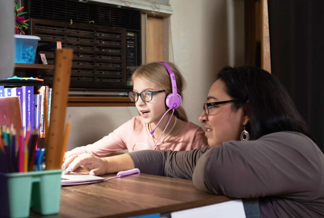 A young girl with glasses and pink headphones focuses on a laptop, guided by an adult woman smiling beside her. The scene is cozy and educational.