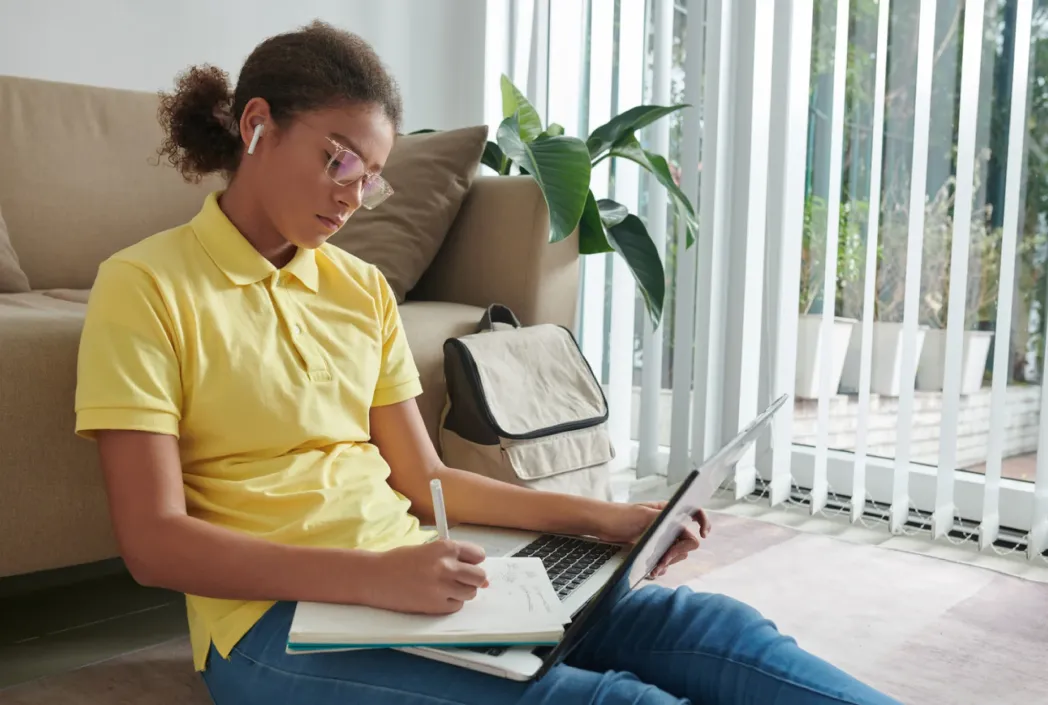 A young person with curly hair and glasses sits on the floor, focused on writing in a notebook. They wear a yellow polo and earphones, with an open laptop nearby.