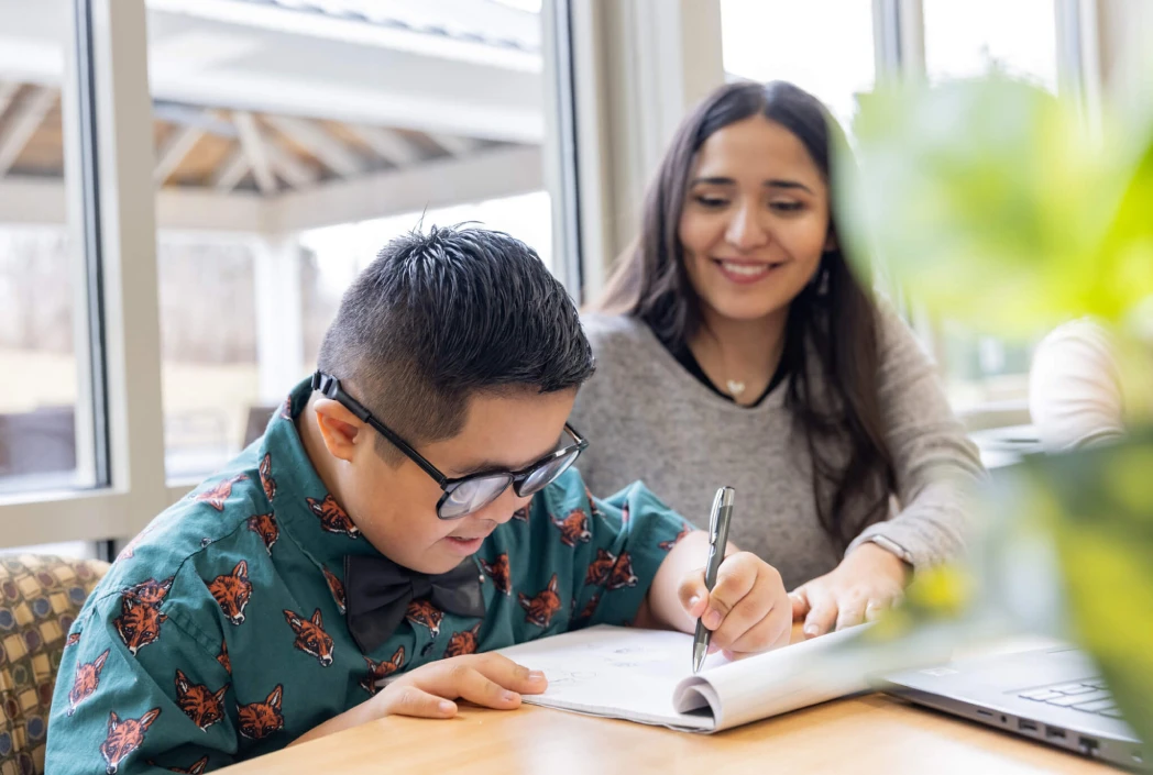 A young boy in glasses focuses on writing in a notebook, while a smiling woman sits beside him, offering support and encouragement. A laptop is visible nearby.