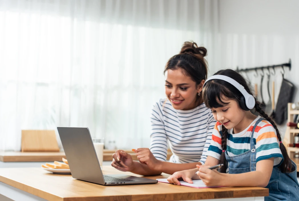 A woman and child sit at a kitchen counter with a laptop open. The woman smiles, guiding the child, who wears headphones and writes in a notebook. Bright, casual setting.