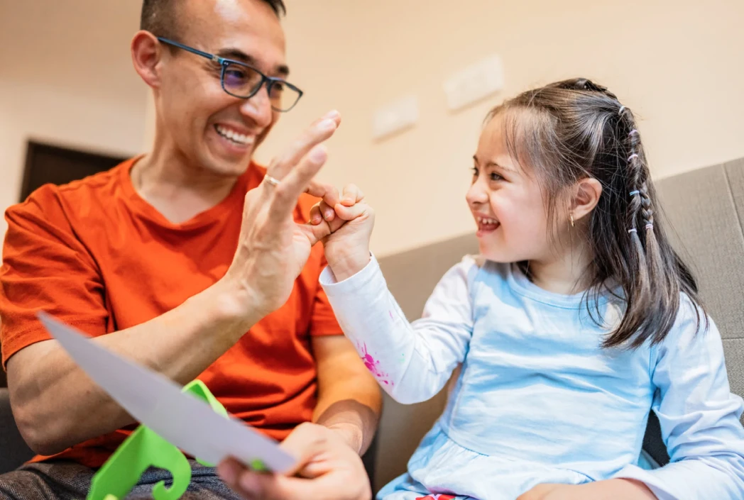 A man in a red shirt and glasses high-fives a smiling girl with Down syndrome in a blue shirt. They sit on a couch, sharing a joyful moment.