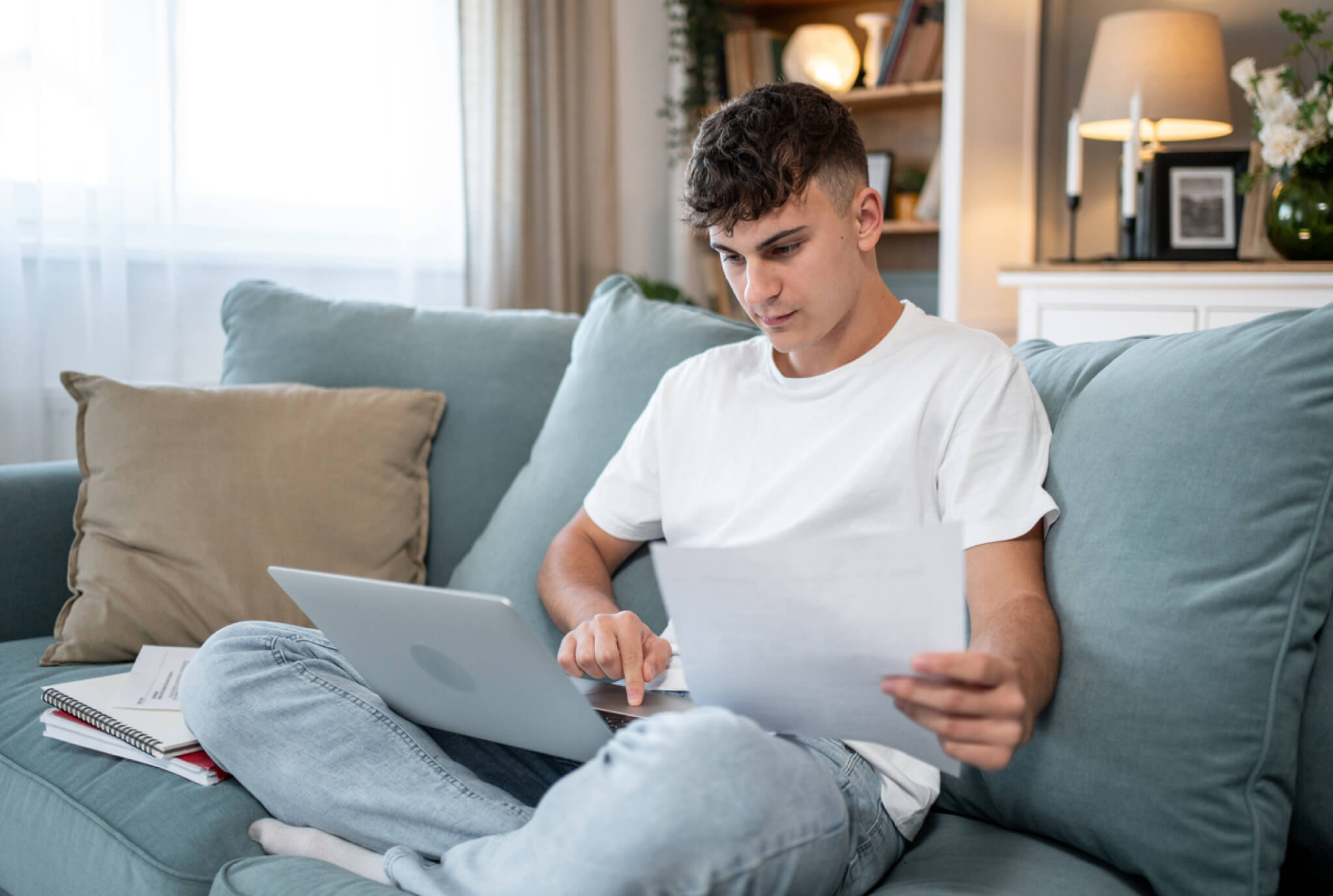 Teenage boy sitting on a couch using a laptop while holding a sheet of paper, with notebooks nearby, focused on studying or completing homework at home.
