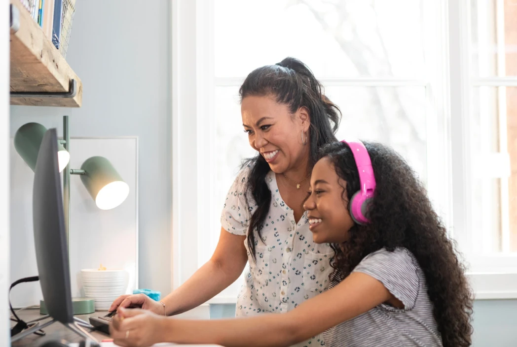 A mother and daughter smile while using a computer at home. The daughter wears pink headphones. The room is well-lit with a cozy, positive atmosphere.
