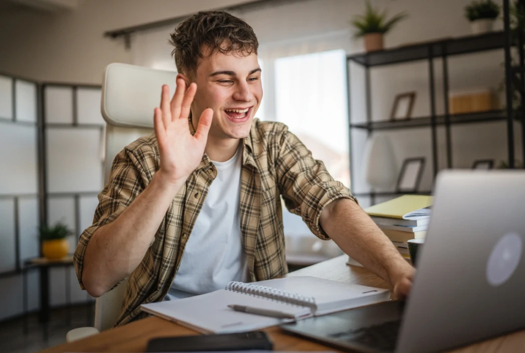 Young man in a plaid shirt smiling and waving at a laptop during a video call. The scene is set in a cozy home office with a warm atmosphere.