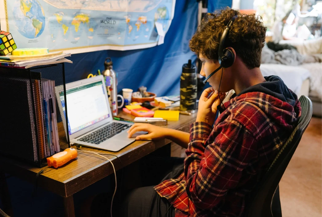 Una persona con auriculares está sentada en un escritorio de madera, enfocada en una computadora portátil con una pantalla abierta. El espacio de trabajo incluye libros, una botella de agua y artículos de papelería, mientras que un mapa del mundo cuelga en la pared azul detrás. Un ambiente acogedor es visible en el fondo.