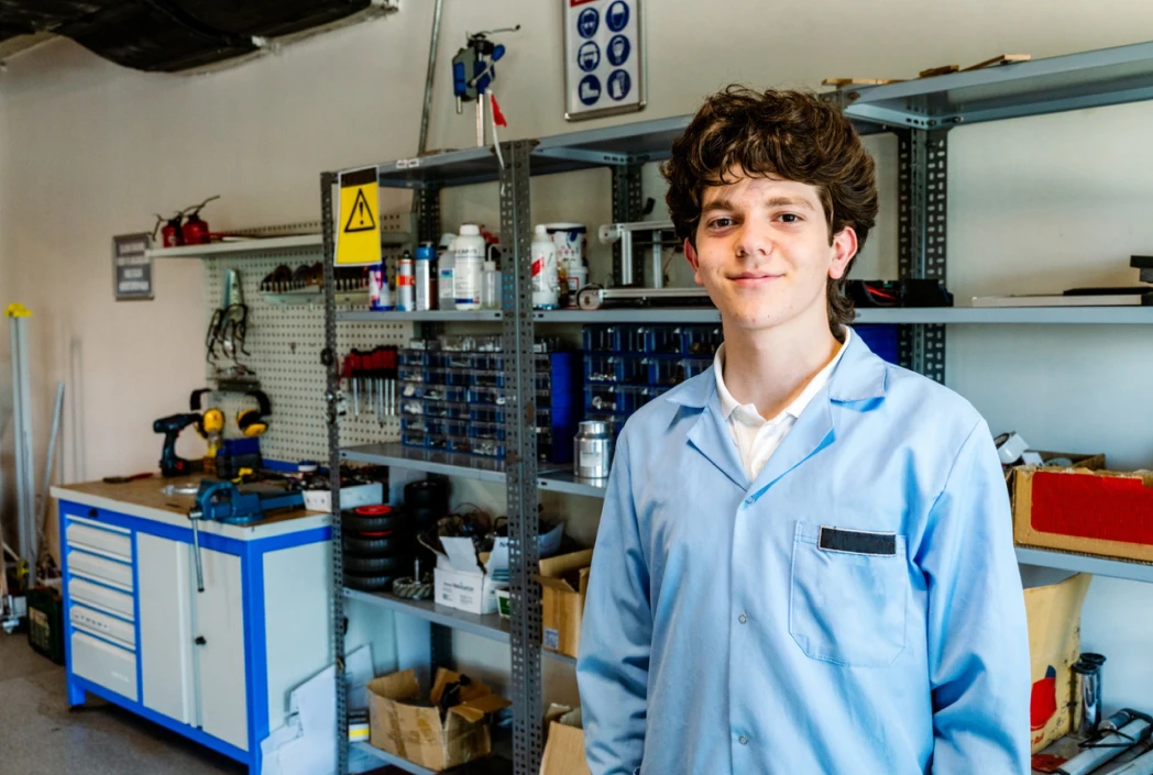 Young person in a blue lab coat stands smiling in a workshop. Behind them are shelves with tools, boxes, and supplies. The space feels organized and industrious.