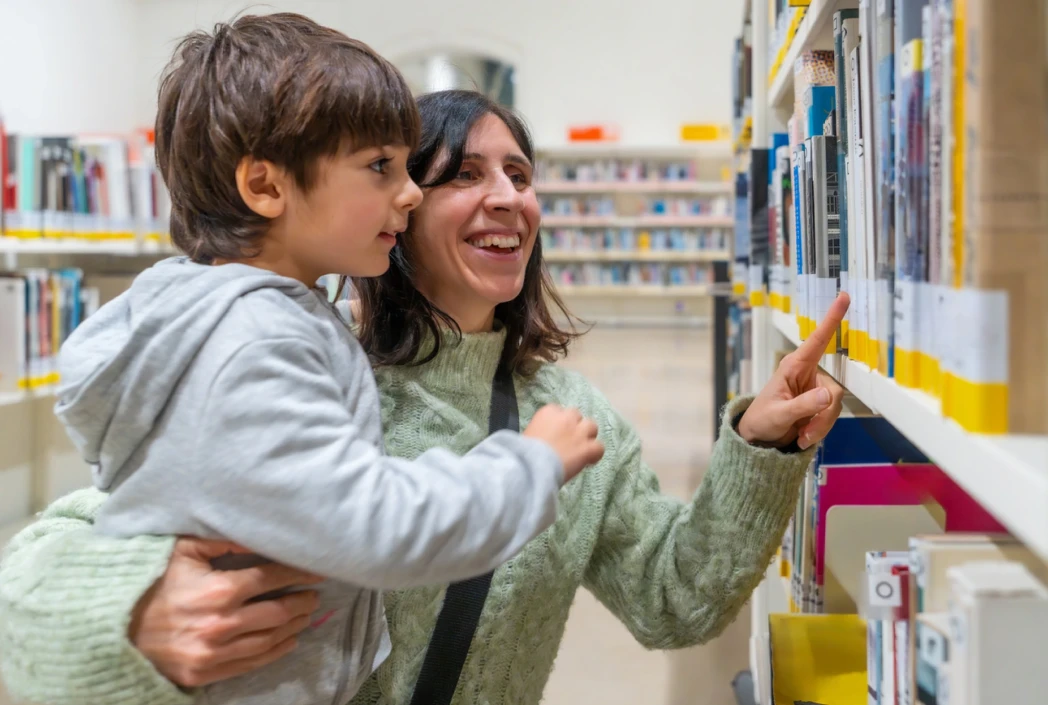 A smiling woman holds a child in a library, pointing at a book on a shelf. The scene conveys warmth and curiosity amidst rows of colorful books.