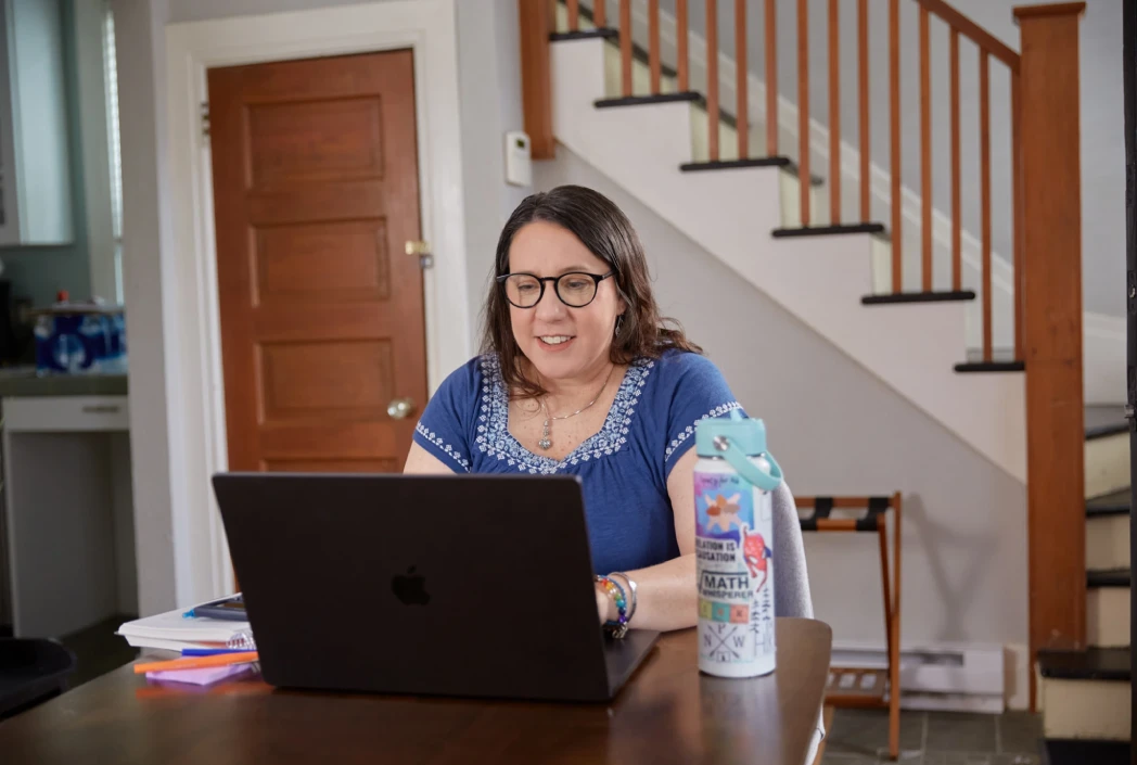 A woman in glasses and a blue blouse sits at a table, smiling at her laptop. A decorated water bottle is beside her, with a staircase in the background.