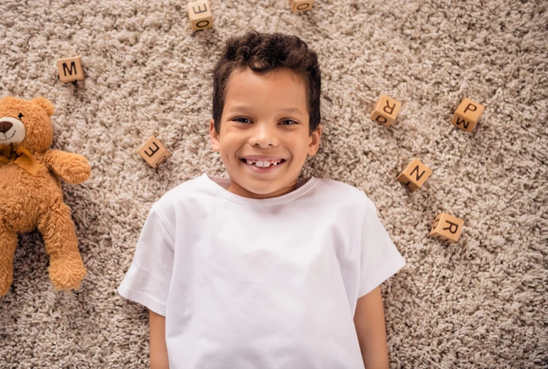 A cheerful child in a white shirt lies on a soft carpet, surrounded by wooden letter blocks and a plush teddy bear, conveying a playful, cozy scene.