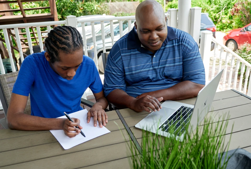 A young person in a blue shirt writes in a notebook, while an older person beside them looks at a laptop. They are sitting outside on a deck.