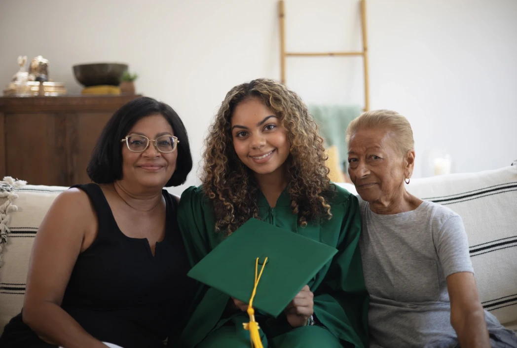 A young woman in a green graduation gown smiling, holds a matching cap, seated with two older women on a couch. The scene is warm and celebratory.