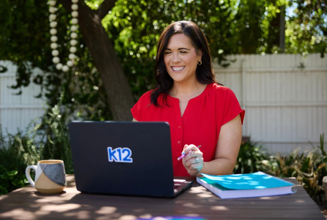 Smiling woman in a red shirt works on a laptop with "k12" logo outdoors, surrounded by greenery. A coffee mug and colorful folders are on the table.