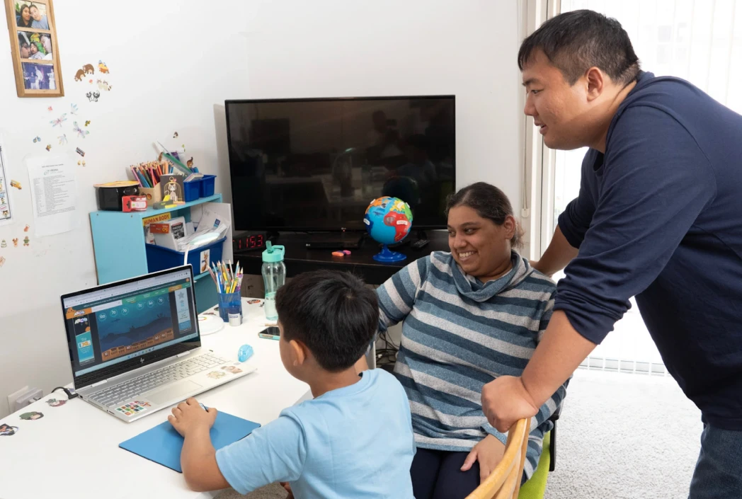 A child using a laptop, playing an educational game, while seated at a desk. Two adults, one smiling, observe and engage, creating a supportive atmosphere.