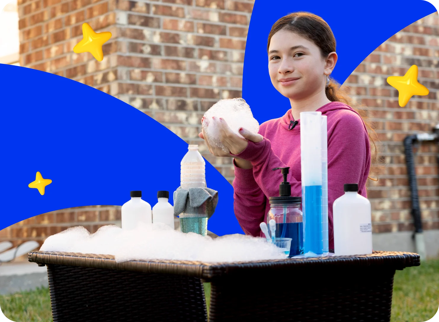 A girl in a pink hoodie holds a large soap bubble, smiling beside a table with bottles and soap. A brick wall and stars on a blue background are behind her.