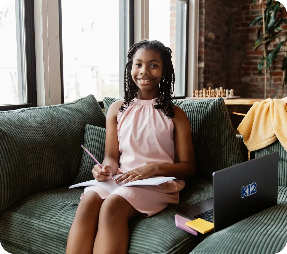 Smiling girl in a pink dress sits on a couch, writing in a notebook. A laptop labeled "K12" is nearby. Bright, cozy room with a chess set.