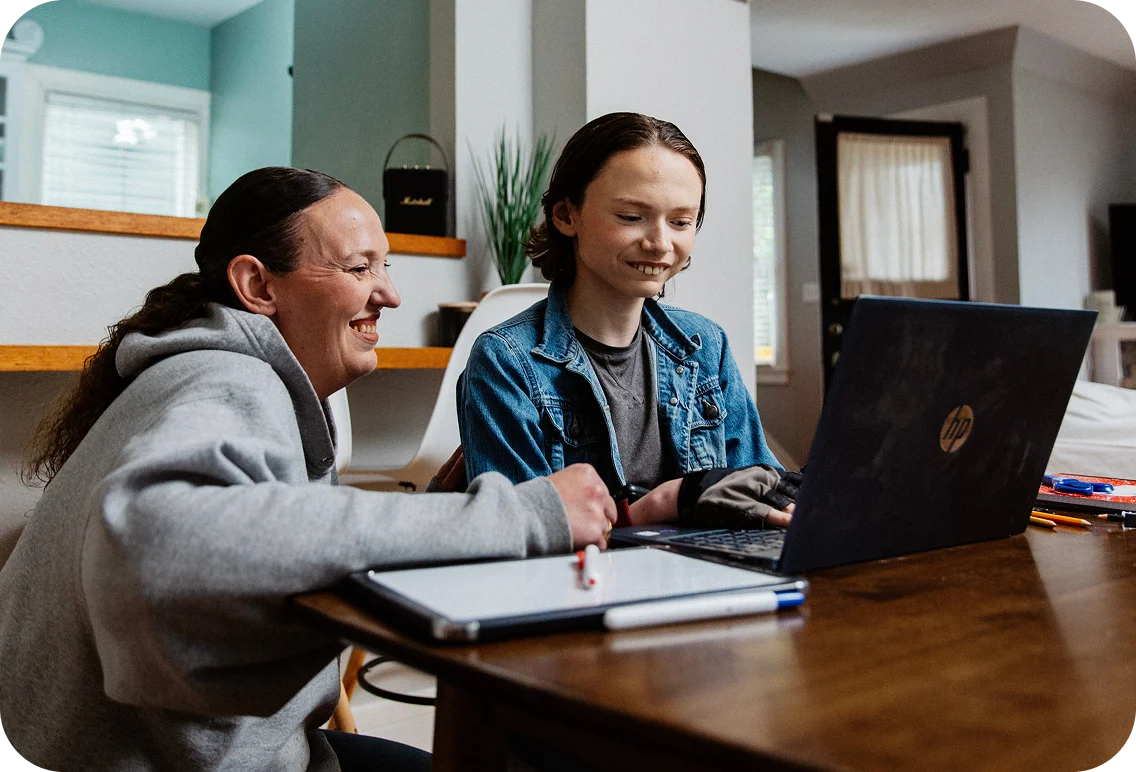 A smiling person and child sit at a wooden table, engaged with an open laptop. The setting is a cozy room with soft lighting, creating a warm atmosphere.