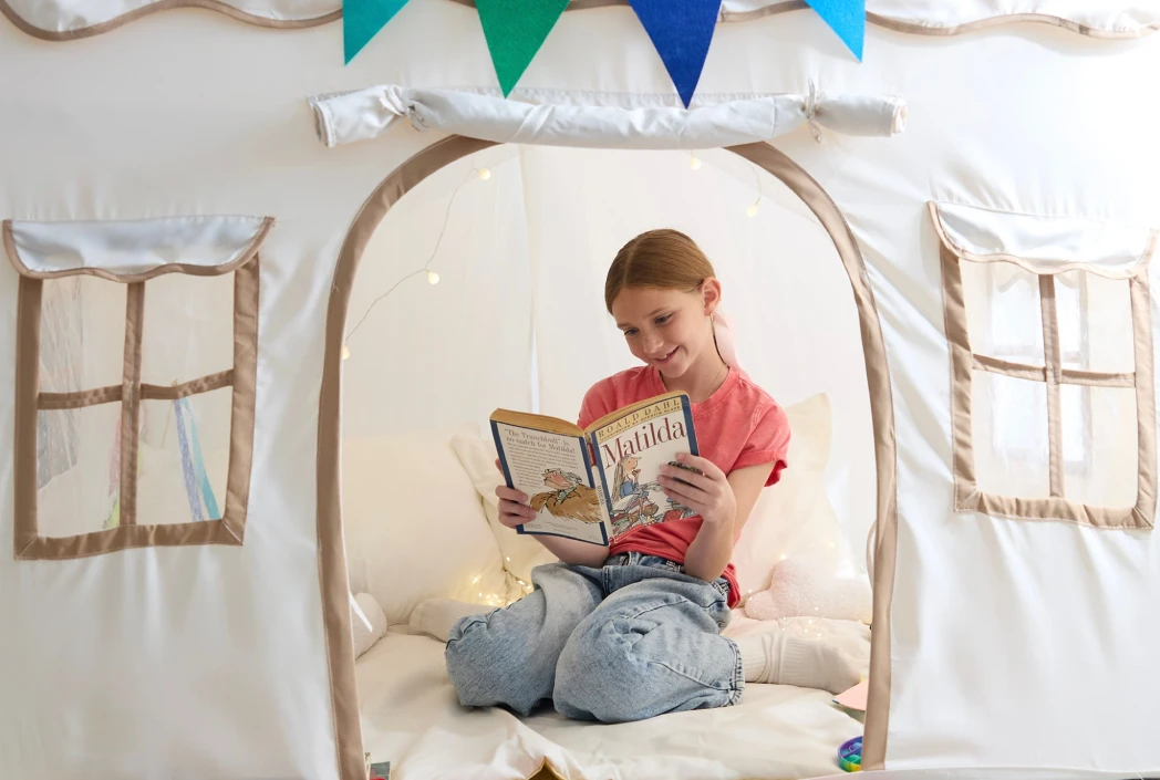 A young girl in a red shirt and jeans sits cross-legged inside a cozy tent reading "Matilda," with string lights hanging, exuding a peaceful and joyful vibe.
